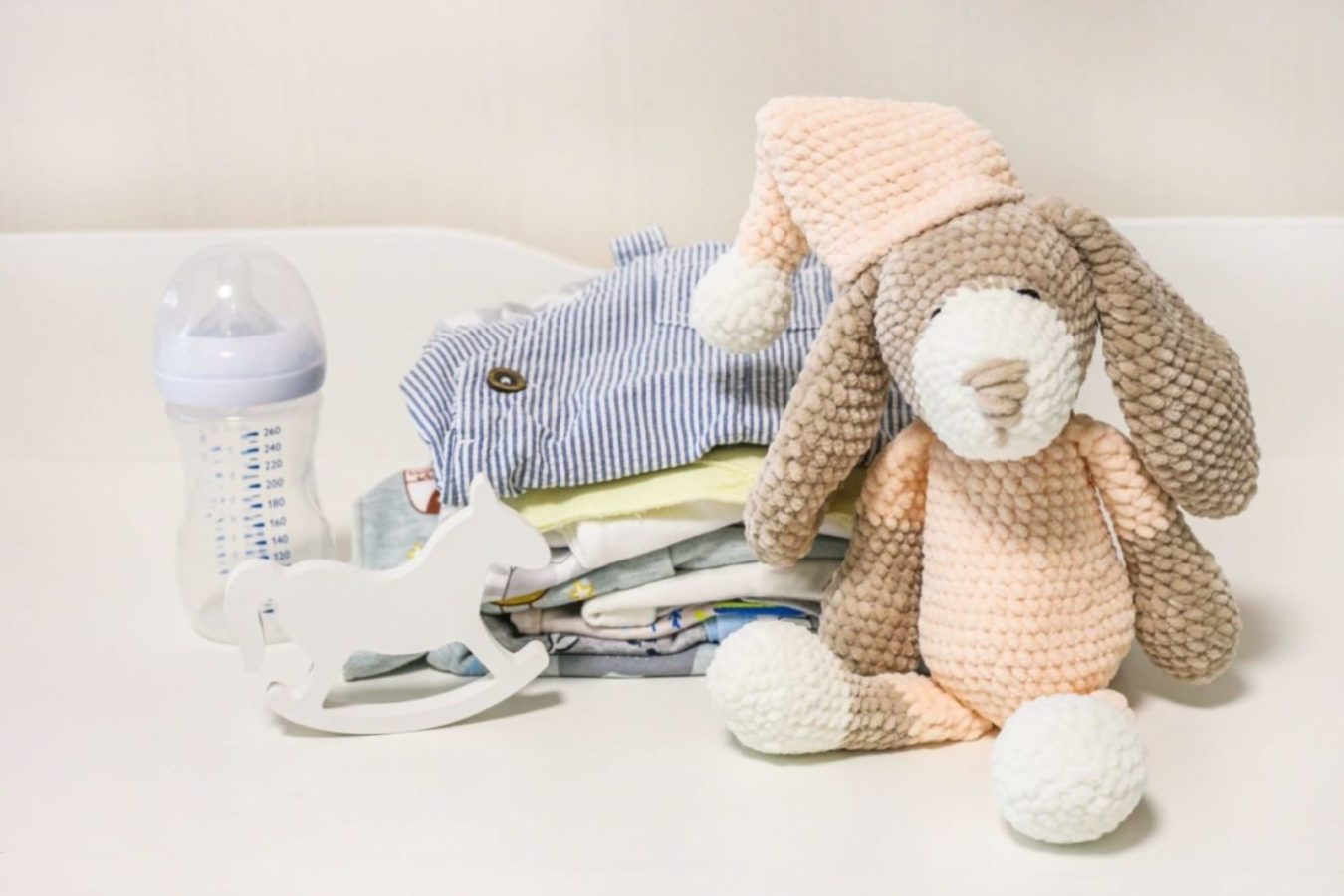 A stack of baby clothes with a stuffed animal, toy horse and baby bottle.